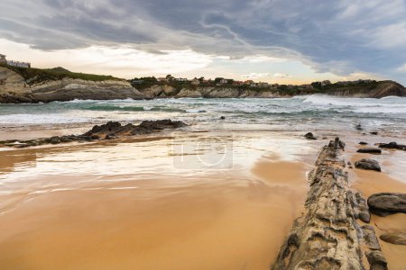 Portio Beach with its golden sand, layers of rock eroded since the Glaciation, the raging Atlantic Ocean, and looming storm clouds. Costa Quebrada Geopark, Cantabria, Spain.