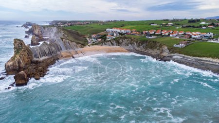 Aerial view of Portio Beach, the coastline with sharp cliffs and rock formations, layers of stone, the stormy Atlantic Ocean, green meadows, and a small village. Costa Quebrada Geopark, Cantabria, Spain.