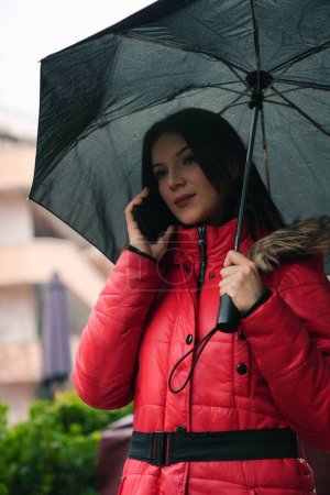 Foto de Una hermosa joven de pie bajo un paraguas y hablando por teléfono. - Imagen libre de derechos
