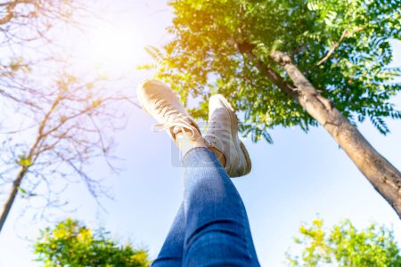 Photo for Legs in white sneakers and jeans against blue sky and green trees - Royalty Free Image