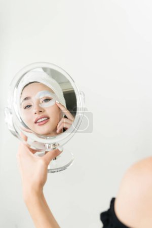 Téléchargez les photos : Miroir reflet de charmante jeune femme avec un bandeau cosmétique collagène - en image libre de droit