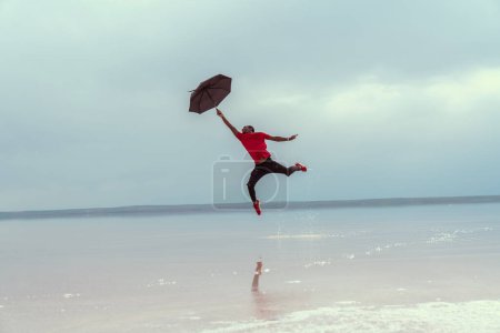 Téléchargez les photos : Portrait d'homme afro-américain avec parapluie sautant au-dessus de la mer - en image libre de droit