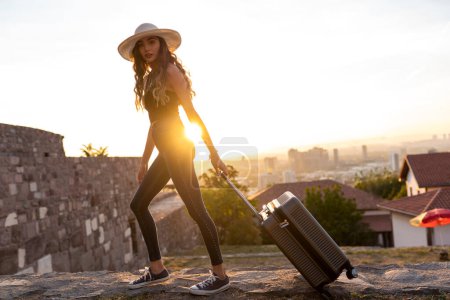 Foto de Mujer joven en sombrero de paja con maleta al atardecer en la ciudad - Imagen libre de derechos
