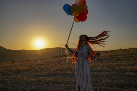 Foto de Joven mujer feliz con globos al atardecer en verano. Mujer feliz corriendo y divirtiéndose con globos en la naturaleza al atardecer - Imagen libre de derechos
