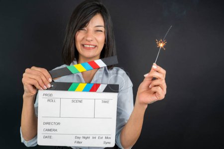 Photo for A happy young woman holding a movie clapboard and a lit sparkler. - Royalty Free Image
