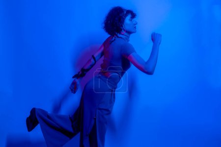 Téléchargez les photos : Haute Femme modèle de mode dans les lumières lumineuses colorées bleu néon et violet posant en studio. Portrait de belle femme avec maquillage rayonnant à la mode. Art design style vif - en image libre de droit