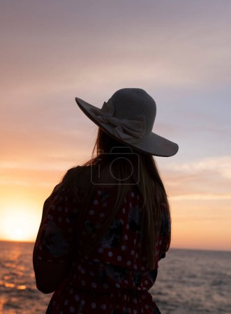 Foto de Una mujer con un sombrero de paja está sentada en una roca cerca del océano. - Imagen libre de derechos