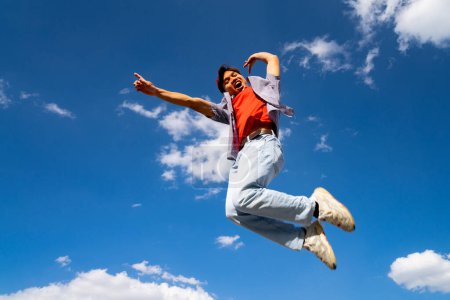 A man is jumping in the air with his arms outstretched. The sky is blue and there are clouds in the background. Scene is joyful and energetic