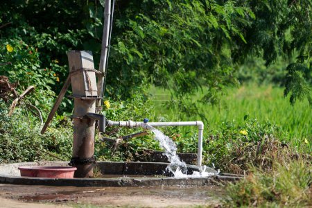 Photo for Water flow from pumping pipe and Old hand operated water pumping rural India high water pressure gushing from plastic pipe of agricultural farm water pump on field background and blue sky with trees and greenery in background. Selective focus - Royalty Free Image