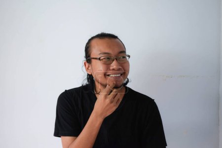 Foto de Hombre asiático adulto vistiendo camiseta negra y gafas con pelo largo Mostrando cara pensante en fondo gris. - Imagen libre de derechos