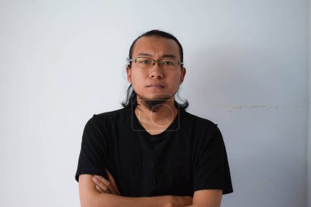Foto de Hombre asiático adulto vistiendo camiseta negra y anteojos con pelo largo Mirando a la cámara con cara seria en fondo gris. - Imagen libre de derechos