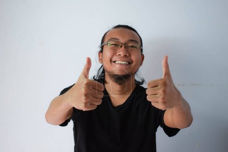 Photo for Adult Asian man wearing black t-shirt with long hair smiling and give thumb up at camera - Royalty Free Image