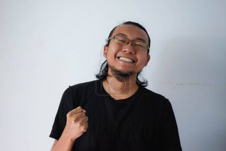 Téléchargez les photos : Homme asiatique adulte portant un t-shirt noir et des lunettes aux cheveux longs Affichant une expression de succès en arrière-plan gris. - en image libre de droit