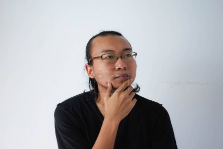 Photo for Adult Asian man wearing black t-shirt and eyeglasses with long hair Showing thinking face in grey background. - Royalty Free Image