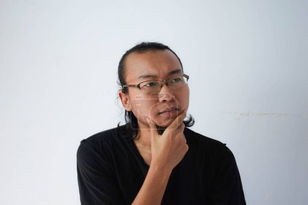 Photo for Adult Asian man wearing black t-shirt and eyeglasses with long hair Showing thinking face in grey background. - Royalty Free Image
