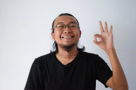 Foto de Hombre asiático adulto con camiseta negra y gafas de vista con pelo largo haciendo bien signo de mano en fondo gris. - Imagen libre de derechos