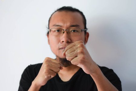 Photo for Adult Asian man wearing black t-shirt and eyeglasses with long hair doing fight pose at camera in grey background. - Royalty Free Image