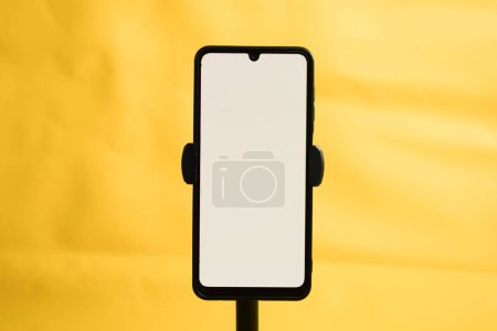 Téléchargez les photos : Portrait téléphone avec écran blanc fixé au trépied sur fond jaune, pour la conception de maquettes. - en image libre de droit