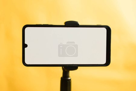 Photo for Landscape phone with white screen fixed to tripod on yellow background, for mockup design. - Royalty Free Image