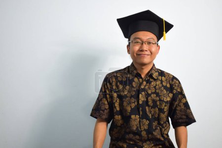Téléchargez les photos : Expressive de l'indonésie adulte mâle porter batik, toga tasse ou diplôme chapeau et lunettes isolées sur fond blanc, expressions de la graduation portrait - en image libre de droit