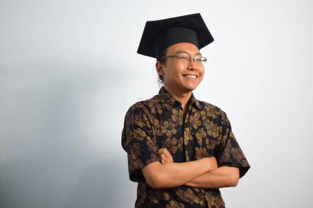 Téléchargez les photos : Expressive de l'indonésie adulte mâle porter batik, toga tasse ou diplôme chapeau et lunettes isolées sur fond blanc, expressions de la graduation portrait - en image libre de droit