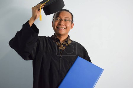 Foto de Expresivo de indonesia adulta ropa de graduación masculina, sombrero y gafas, graduación masculina asiática traer certificado azul en blanco aislado sobre fondo blanco, expresiones de graduación de retrato - Imagen libre de derechos