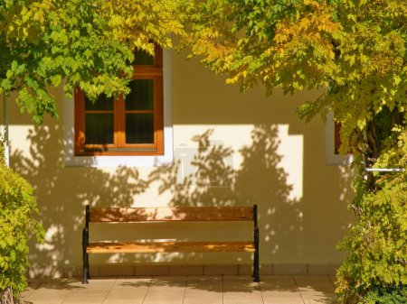 Autumn atmosphere, a house with an ancient window and a bench. Shadow play on the wall and yellowing leaves on the trees.Piestany spa, Slovakia.
