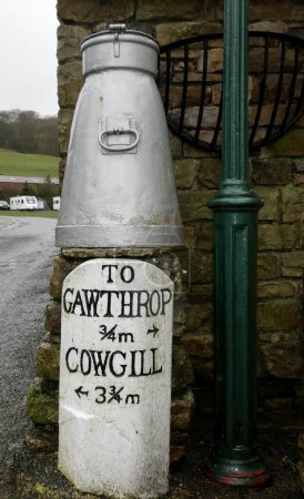 Photo for Milk Churn over Road sign to Gawthrop and Cowgill. Green lamppost alongside. Dent, Cumbria, UK. February 15, 2020. - Royalty Free Image