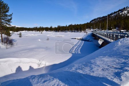 Foto de Road Bridge over snowed over frozen river, Aust-Agder, Norway. Trees behind. Blue skies. Shadows on the snow. Feb 2023. - Imagen libre de derechos