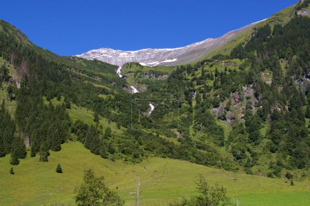 Baumbestandener Hang mit Wasserfall und schneebedeckten Bergen dahinter, unter blauem Himmel. Großglockner, Österreich.