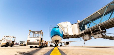 Photo for Blue aircraft parked on tarmac with gate attached to front door.Bagage handling is standing by. Bright Blue sky. Yellow guidance line painted on concrete. High quality photo - Royalty Free Image