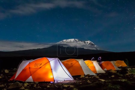 Orange tents illuminated from inside before Mount Kilimanjaro during nighttime. Stars and Milky Way visible. High quality photo