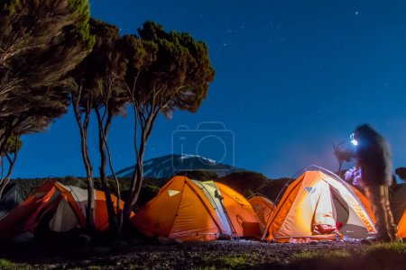 Orange tents illuminated from inside before Mount Kilimanjaro during nighttime. Stars and Milky Way visible. High quality photo