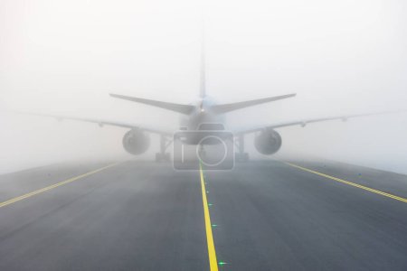 Airplane taxing on taxiway in white fog conditions. Bright yellow taxi line on black tarmac. Aircraft seen from straight behind. High quality photo