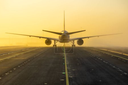 Photo for Airplane taxing on taxiway in orange morning fog conditions. Bright yellow taxi line on black tarmac. Aircraft seen from straight behind. High quality photo - Royalty Free Image