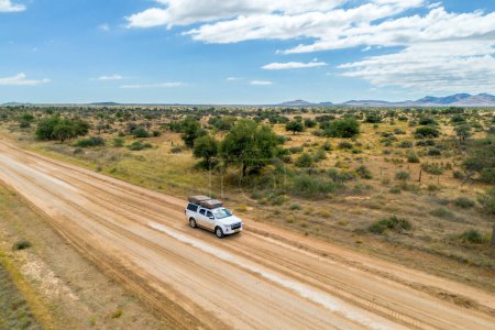 Foto de Drone imagen aérea de vehículo todoterreno conduciendo en un camino de tierra en el arbusto africano. Carretera de tierra polvorienta amarilla y naranja, arbustos verdes y cielo azul. Namibia. Foto de alta calidad y alta resolución - Imagen libre de derechos