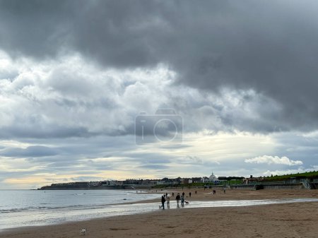 Foto de Nubes de lluvia dramáticas oscuras sobre el oleaje en la playa con arena dorada y olas en Whitley Bay, Newcastle, Inglaterra. Tiempo típico inglés con lluvia. Gente paseando al perro en la playa. - Imagen libre de derechos