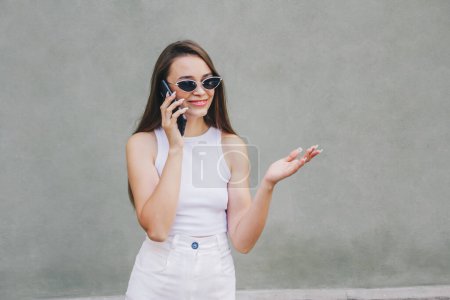 Photo for Beautiful european girl with long hair in stylish sunglasses on the beige background talks by phone and articulates with her hand. Banner, place for text concept. - Royalty Free Image