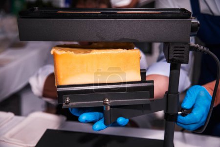 Electric Cheese Melter Slicing through a Wheel of Raclette Cheese