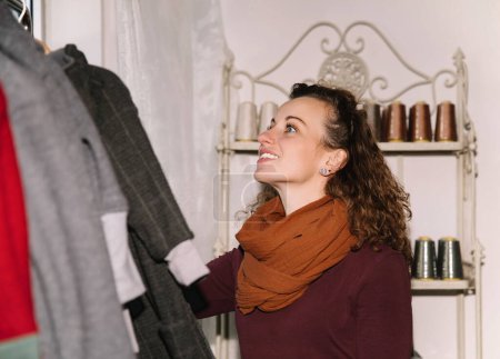 A woman with curly hair and a warm scarf happily browsing through a selection of clothes in a boutique, surrounded by elegant interior decorations