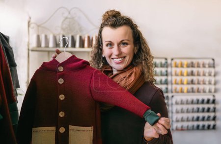 Una mujer sonriente con el pelo rizado sostiene una chaqueta roja en una pintoresca boutique, concepto de tienda