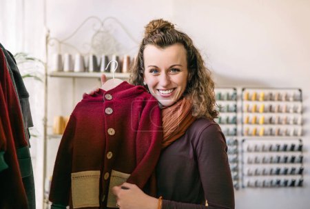 Eine lächelnde Frau mit lockigem Haar hält eine rote Jacke in einer urigen Boutique, umgeben von verschiedenen bunten Fäden und Modeaccessoires