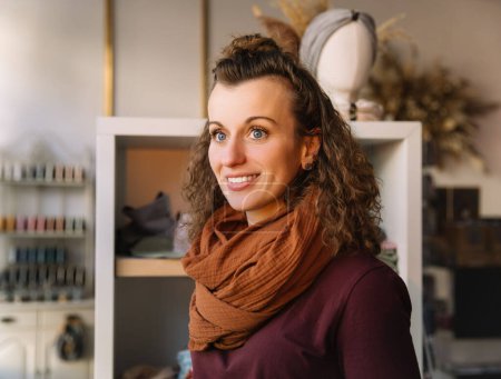 Confident curly-haired woman smiling gently in a well-lit craft shop, surrounded by creative materials and elegant decor