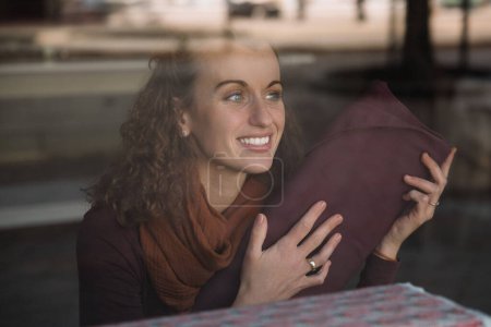 Curly-haired woman seen through a glass storefront, holding a piece of burgundy fabric and smiling warmly, Store concept