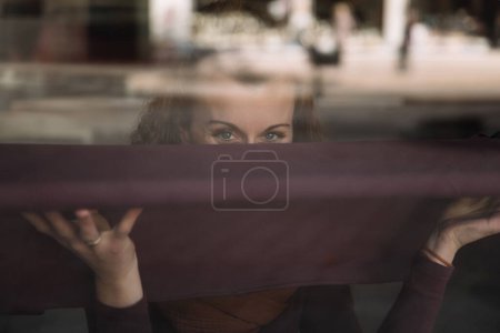 Curious woman with expressive eyes looking through a reflective glass window, hands pressed against the surface, urban background softly blurred