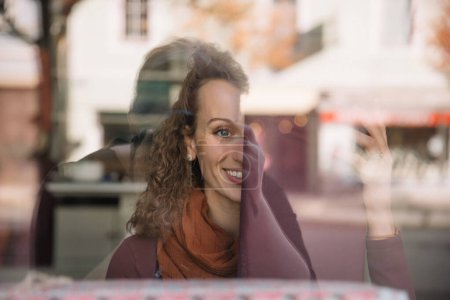 Lächelnde Frau mit lockigem Haar genießt den sonnigen Tagesblick hinter einem Glasfenster, das städtische Leben spiegelt sich um sie herum