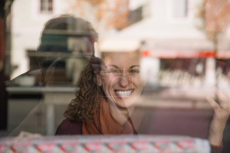 Mujer joven radiante sonriendo ampliamente detrás de una ventana reflectante, café urbano borrosa en el fondo con tonos otoñales