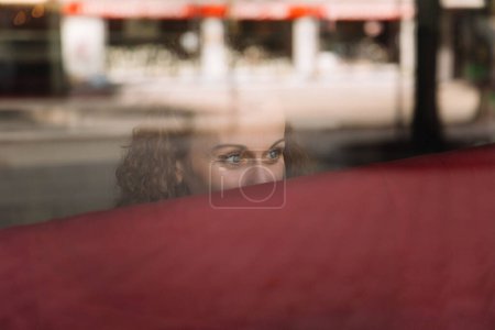 Nachdenkliche Frau mit lockigem Haar, die hinter einem spiegelnden Glasfenster hervorblickt, Stadtleben sichtbar im weichen Fokushintergrund