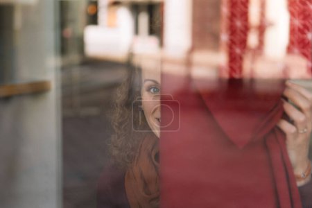 Femme gaie avec les cheveux bouclés regardant à travers une fenêtre en verre, sourire lumineux visible, environnement urbain doucement concentré en arrière-plan