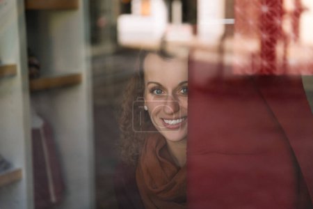 Sourire captivant d'une jeune femme aux cheveux bouclés vu à travers une fenêtre de café, lumière du jour chaude illuminant son visage